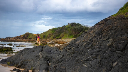 Fototapeta na wymiar Beautiful girl stand on large rocks and admires powerful waves in popular Byron Bay, NSW, Australia