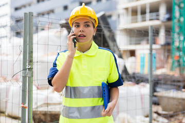 Positive female builder in hardhat standing with folder and discussing project by the phone near...