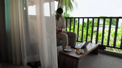 Young adult caucasian holiday maker is surfing internet on smartphone sitting on hotel room balcony in front of open laptop. He is checking messages and preparing for distant work. Freelance in trip - Powered by Adobe