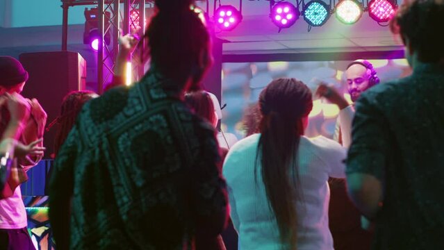 Happy woman doing karaoke at party, singing songs with DJ in front of friends on dance floor. Confident girl having fun with live show perfromance next to electronic mixing station at nightclub.