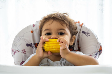 Baby eating corn with funny expression in a high chair. Mention of attention to children's nutrition, food waste and differences in eating capacity according to economic class.