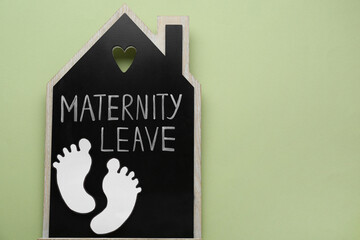 Wooden house figure with words Maternity Leave and paper cutout of baby feet on pale green background, top view. Space for text