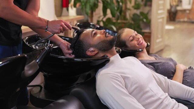 A cheerful Indian man with his pregnant wife in a hair salon getting his hair washed while leaning his head in a shampoo bowl. High-quality 4k footage