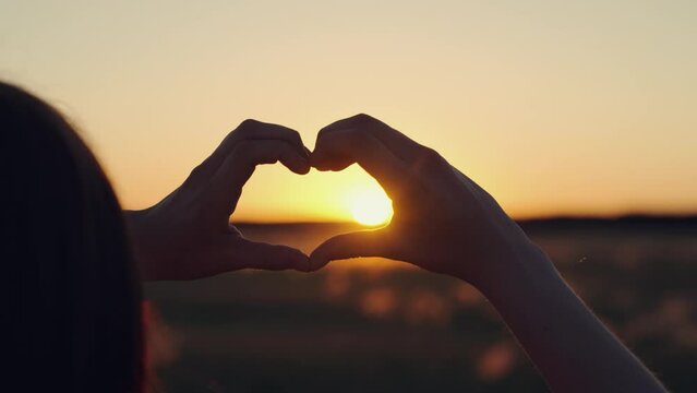 Heart shape made by fingers of teenager against background of the sun. Chidhood dream. Silhouette of child of girl in park at sunset shows symbol of heart with her fingers. Concept of health, love,