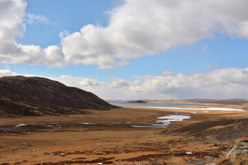 A wide bed of a frozen shallow river flows into a wide lake on a sunny autumn day, surrounded by snow-covered mountain ranges.