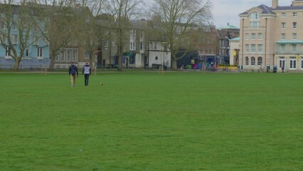 Friends walk across grassy field with dog by colorful homes on cloudy day - Powered by Adobe