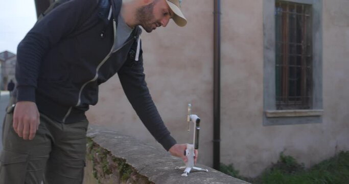 Male Tourist Wearing Backpack Extending Tripod Legs On Mobile Gimbal And Placing It On Low Stone Wall