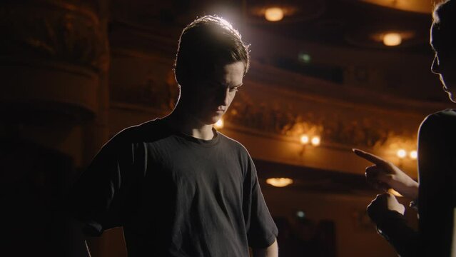 Concentrated ballet male dancer stands with ballerina on theater stage and discusses performance. Couple of ballet dancers on rehearsal before start of a show. Illuminated theatrical hall. Dolly zoom.