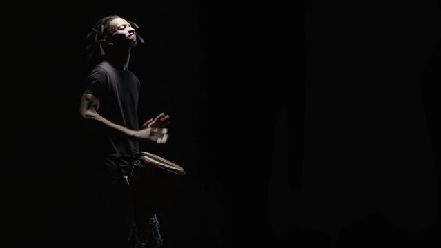 Black musician plays standing the wooden hand drum in a dark studio fast and passionately. Music performer vigorously beating with hands the traditional djembe between his legs