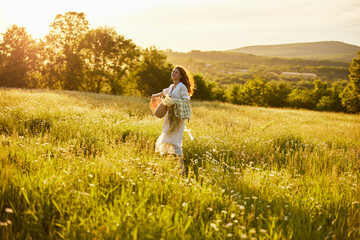 a woman in a white dress with a basket of daisies and a straw hat stands in a field during sunset in a light breeze