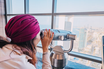 Girl tourist at the observation viewpoint using binoculars and looking to Abu Dhabi city harbor with yachts and cruise ships
