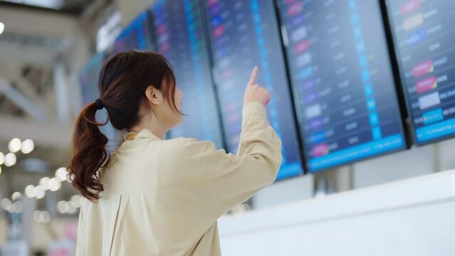 Young asian woman with passport and boarding pass as a hand in international airport looking at the flight information board, checking her flight