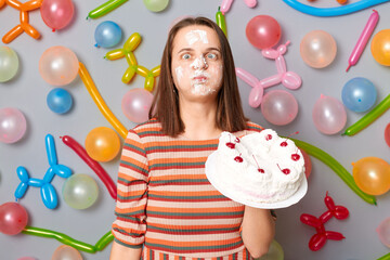 Obraz na płótnie Canvas Funny crazy woman in striped dress standing against gray wall decorated with colorful balloons birthday girl with cake cream on face crossing eyes.