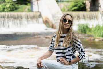 Fototapeta na wymiar A woman in sunglasses, a striped top and white jeans near the river against the backdrop of the dam