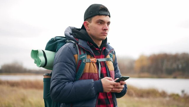 Portrait of young guy tourist backpacker in cap with smartphone finds route on map hiking in autumn. He looks around in search of right path. Hike, tourism, travel, explore wild nature concept.
