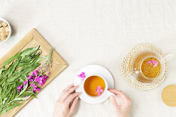 Female hands hold cup with fireweed tea, transparent glass teapot, herbal hot tea from green leaves of ivan chai on tablecloth. Top view healthy drink and wild flowering willow-herb, tea time