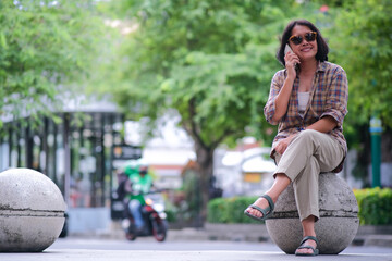 Asian woman sitting alone on a round stone seat in the park, making a phone call