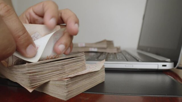 Select focus of hands counting a lot of thousands Thai baht banknotes money near laptop or notebook computer. The baht is the official currency of Thailand.