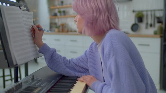 Close-up of inspired talented pretty pink haired female musician in creative process composing music using electronic keyboard, writing musical notes into lead sheet on sheet music stand. Side view.