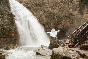Happy woman in a white dress stands on a stone with a waterfall behind