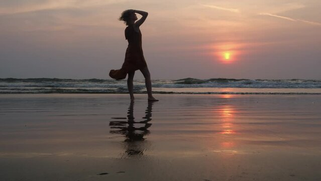 Back view of happy woman standing on the seashore, admiring the beautiful sunset, enjoying vacation and freedom. Female silhouette in a long fluttering dress is reflected in a water mirror.
