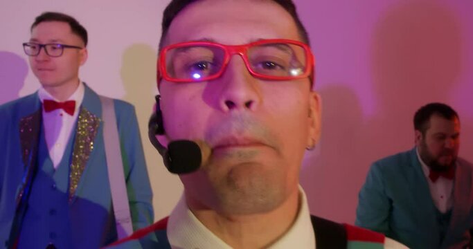 Portrait of man in red glasses musician singing rap in headset microphone performing with band, playing musical instruments and dancing on performance, live concert in club looking at camera.