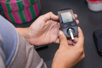 Young Latino man doing blood sugar test in the kitchen. Blood glucose test. Drawing blood for medical test. Using glucose meter in the kitchen.