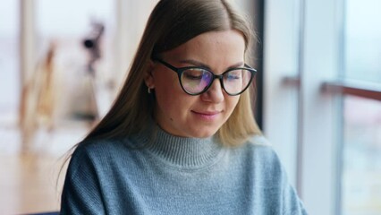 Calm positive Caucasian woman wearing glasses sits at desk. Lady stops typing on her laptop, takes off glasses and smiles to camera.