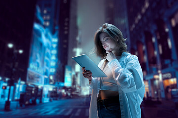 Young pretty girl standing on street at night and looking on tablet with doubtful, thoughtful face. Blurred city background. Online shopping. Modern technologies, youth, communication and business