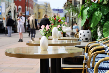 Street cafe in city with empty tables outdoor on walking people background. Flowers in vases on round tables in sunny day