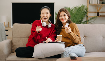 Obraz premium Two asian beauty smiling young women sitting on sofa Attractive casual girl feel happy and relax,having fun watch comedy video on TV in house. Activity lifestyles concept.