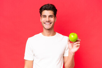 Young caucasian man with an apple isolated on red background smiling a lot