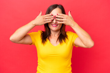 Fototapeta premium Middle-aged caucasian woman isolated on red background covering eyes by hands and smiling