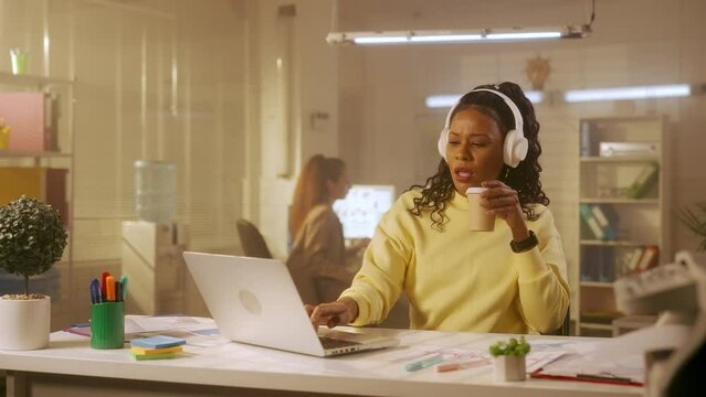 A young African American woman is typing on a laptop keyboard, drinking coffee and enjoying music on headphones. Black female programmer or designer in a yellow sweatshirt sits at a table in office.