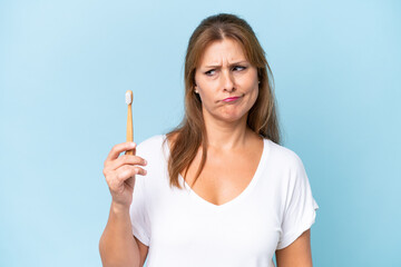 Middle-aged caucasian woman brushing teeth isolated on blue background with sad expression