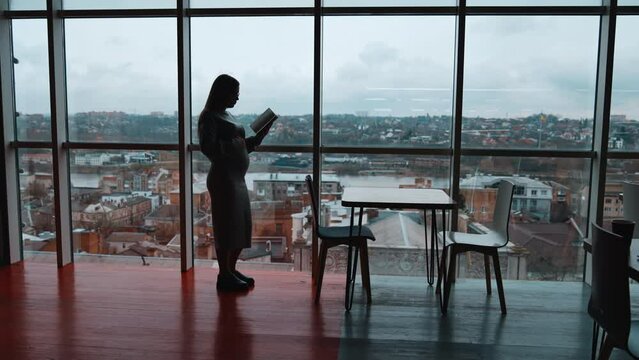 Silhouette of pregnant woman standing near the big panoramic window. Expecting lady reads a book touching her belly. Suddenly woman feels bad and bends holding by the chair.