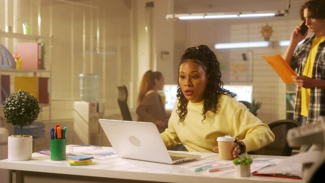 A young African American woman late for work runs into the office, sits at the table and starts typing on the laptop keyboard. Black female in a yellow sweatshirt sits at a table in the office.