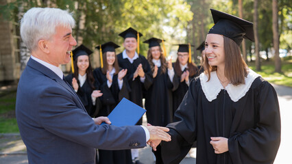 The teacher shakes hands with the student and presents the diploma outdoors. A group of university graduates.