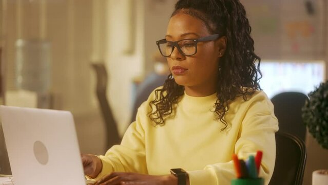 A young African American woman is typing on a laptop keyboard, looking at the camera and smiling. A black female in a yellow sweatshirt sits at a table in the office. Close up.