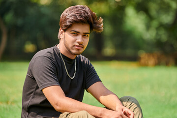 Attractive young indian man portrait in black t shirt and silver neck chain sitting on green lawn...