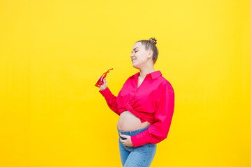 Happy pregnant woman with a smile on her face. Cheerful pretty pregnant woman in a pink shirt and pink glasses on a yellow background. Young bright pregnant woman.