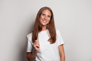 Fototapeta premium Good-looking pretty smiling lady in white t-shirt touching her hand her hair and looking at camera on white
