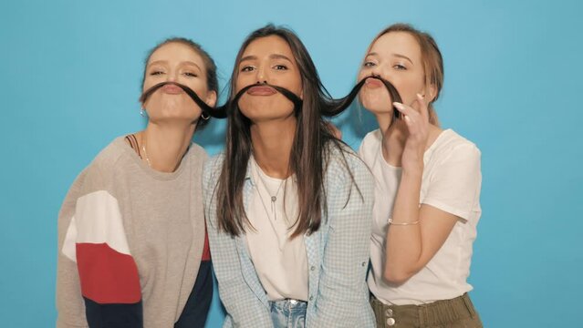 Three young beautiful smiling hipster girls in summer clothes. Sexy carefree women having fun.Positive models going crazy near blue wall in studio.They make moustache with hair