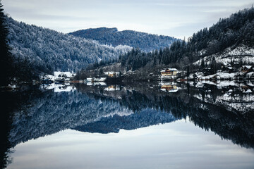 Fototapeta premium Houses on the lakeside against a mountain covered with trees in winter with reflection in water