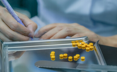 Yellow sugar coated tablet pill on stainless steel drug tray. Pharmacist hand holding pen and...