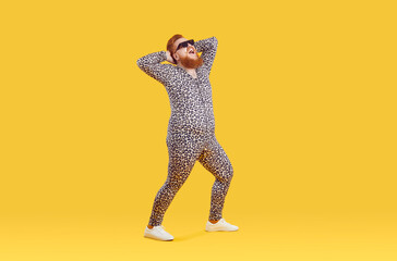 Funny cheerful fat guy in crazy animal print PJs having fun in modern fashion studio. Happy excited fat man wearing comfortable leopard pajamas and sunglasses posing isolated on yellow background