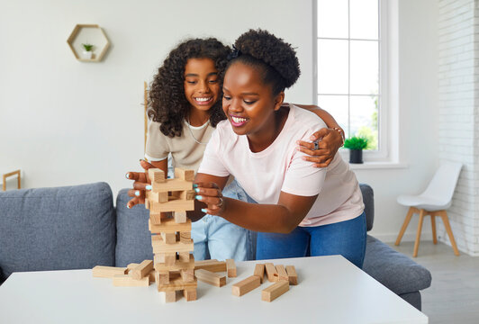 Happy young family enjoy board game at home. Cheerful, joyful, smiling Afro American mother and child daughter standing by table in living room, playing game and putting wooden block on top of tower