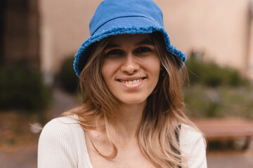 Pretty young caucasian woman looking at camera, spends her leisure time on walk in street. Brunette wears blue bright panama hat, top with long sleeves. Girl look at camera.