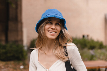 Pretty young caucasian woman looking away spends her leisure time on walk in street. Brunette wears blue bright panama hat, top with long sleeves. Positive emotions concept.