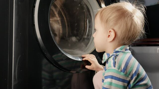 Curious little baby looking at spinning drum of washing machine at home. Doing housework and chores, children education and development